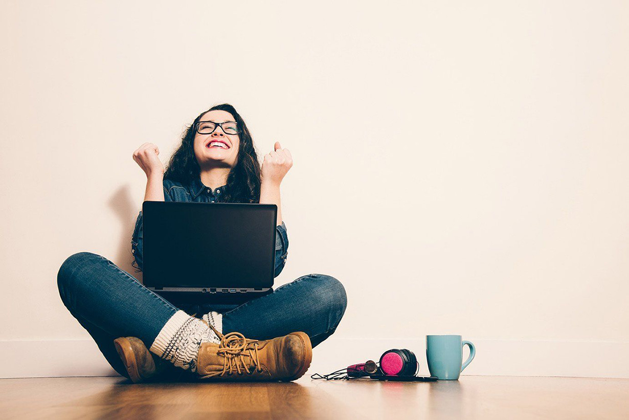 A young woman, arms up in celebration, sitting on the floor with a laptop.